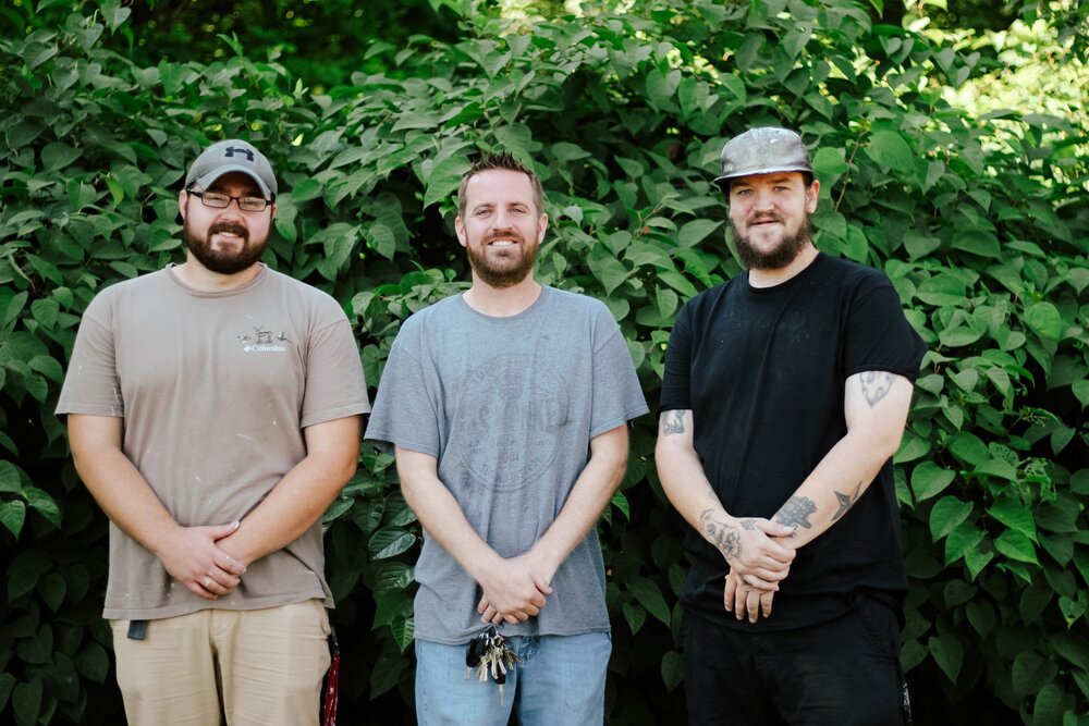 Three men smiling and standing next to each other with leaves in the background